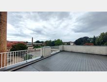 Eine Dachterrasse mit grauem Holzboden und weißem Geländer, die eine Aussicht auf umliegende Gebäude, Bäume und einen Backsteinschornstein bietet, unter einem bewölkten Himmel. Im Hintergrund sind mehrere rote Dächer und eine grüne Baumlandschaft erkennbar.