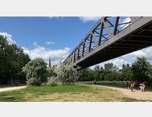 Eine stählerne Fußgängerbrücke überspannt eine grüne Parklandschaft mit Bäumen und Wiesen, im Hintergrund ist der spitze Turm einer roten Backsteinkirche sichtbar. Im Vordergrund gehen Personen auf einem Weg neben einer Sandfläche.