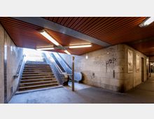 Unterirdischer Zugang mit breiter Treppe, daneben Rolltreppen, die aus dem U-Bahn-Bereich nach oben führen. Die Wände sind aus grauem Beton, einige davon mit Graffiti, und die Decke hat holzähnliche Verkleidungen mit leuchtenden Deckenlampen.