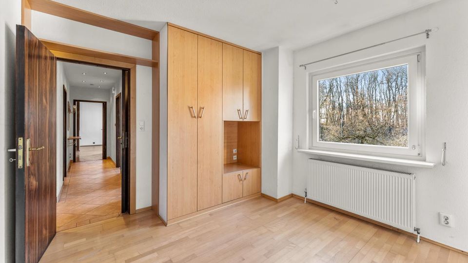 Heller, leerer Wohnraum mit hellem Parkettboden, eingebautem Holzschrank und offener Tür zum gefliesten Flur. Rechts ein großes Fenster mit Blick auf Bäume sowie ein Heizkörper darunter.