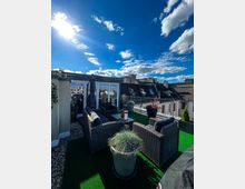 Dachterrasse mit Kunstrasen, mehreren dunklen Rattanmöbeln und Pflanzen in Blumentöpfen; im Hintergrund sind umliegende Gebäude unter einem sonnigen, wolkigen Himmel sichtbar.