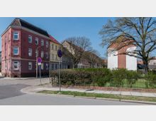 Straßenszene in einem Wohngebiet mit alten, mehrstöckigen Häusern in roten und gelben Fassadenfarben, einem gepflegten Garten mit Hecken und einem großen Baum im Vordergrund. Im Hintergrund sind gepflasterte Gehwege, Straßenschilder und weitere Gebäude sichtbar.