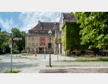 Straßenecke mit Kopfsteinpflaster, Straßenschildern für die "Wasgenstraße" und "Tewsstraße" sowie einem Gebäude im Hintergrund mit rotem Banner und Rankenbewuchs. Rechts sind Bäume und Büsche sichtbar, und eine orangefarbene Mülltonne steht auf dem Vorplatz.