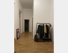 Ein Flur in einer Wohnung mit hellem Holzfußboden, weißen Wänden und hohen Decken. Rechts steht eine Garderobe mit hängenden Jacken und Schuhen darunter, links lehnt ein gerahmtes Poster an der Wand, und im Hintergrund sind Türen zu weiteren Räumen sichtbar.