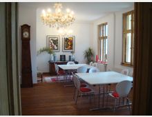 Ein Seminarraum mit Holzboden, weißen Wänden und großen Fenstern mit Holzrahmen. Der Raum ist mit weißen Tischen, roten und weißen Stühlen, einem Kronleuchter, einer antiken Standuhr, einer Kommode und abstrakter Wandkunst ausgestattet, ergänzt durch grüne Pflanzen.