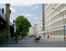 Städtische Straßenszene mit einer modernen Gebäudefassade rechts, einem erhöhten U-Bahn-Viadukt aus Stein links und einer Reihe grüner Bäume entlang des Gehwegs. Im Vordergrund fährt ein Radfahrer eine breite Straße hinüber, während Autos und ein Bus im Hintergrund sichtbar sind.