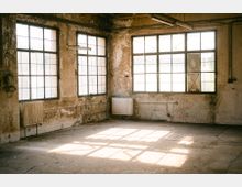 Ein leerer, verlassener Raum mit großen rechteckigen Fenstern, die Tageslicht hereinlassen. Die Wände sind abgenutzt und haben sichtbare Stellen von Verfall, während Heizkörper und freiliegende Rohre an den Wänden angebracht sind.