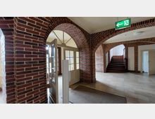 Eingangsbereich eines Gebäudes mit gemauerten Rundbögen und roten Backsteinwänden. Zu sehen sind eine offene Holztür mit Glaseinsätzen, ein Treppenaufgang mit Holzstufen rechts, beige Fliesenboden und ein Notausgangsschild an der Decke.