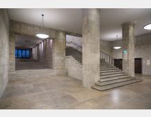 Heller Treppenhausbereich in einem Gebäude mit Wänden, Säulen und Böden aus hellbeigem Stein. Zwei Treppen mit Metallgeländern führen zu unterschiedlichen Ebenen, und große, runde Deckenleuchten beleuchten den Raum.