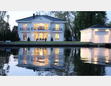 Zweistöckige, beleuchtete Villa im klassischen Stil mit Säulen und Balkonen, gelegen an einem ruhigen Gewässer. Rechts ein separates, ebenfalls beleuchtetes Garagengebäude; umliegende Bäume und eine gepflegte Rasenfläche ergänzen die Szene.