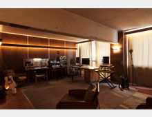 Innenraum eines Tonstudios mit Holzvertäfelung und warmen Wandleuchten, ausgestattet mit Mischpulten, Aufnahmegeräten und Monitoren an mehreren Arbeitsplätzen. Große, mit Vorhängen bedeckte Fenster sowie Musikinstrumente wie Gitarren, Keyboard und Mikrofonständer prägen den Raum.