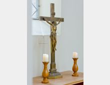 Bronzenes Kruzifix auf einem Holztisch, flankiert von zwei brennenden Kerzen auf Holzleuchtern; im Hintergrund ein Fenster mit weißem Rahmen.