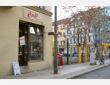 Straßenszene mit einem kleinen Café an einer beigefarbenen Hausfassade, mit einem Schild „Café & more...“. Vor dem Eingang steht ein Werbeschild, daneben ein roter Getränkekühlschrank. Im Hintergrund sind ein Zaun, Bäume ohne Laub, ein Fahrrad und mehrstöckige Gebäude zu sehen.