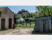 Ein Hof mit zwei alten, verwitterten Schuppen, einem Müllcontainer und einem Metallzaun, der Zugang zu einem kleinen Garten mit Gewächshaus gibt. Im Hintergrund sind weitere Gärten, Bäume und mehrere Häuser mit roten Ziegeldächern zu sehen.