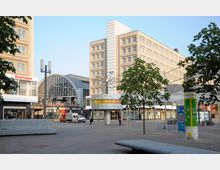 Blick auf den Alexanderplatz in Berlin mit der Weltzeituhr im Vordergrund, flankiert von Bäumen, einer modernen Glasfassade und umliegenden Gebäuden. Im Hintergrund ist der Bahnhof Alexanderplatz mit seinem charakteristischen Bogendach zu sehen.