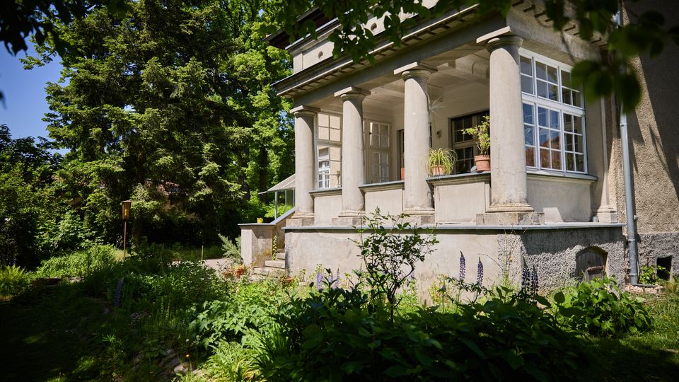 Seitenansicht einer älteren Villa mit überdachter Veranda, getragen von mehreren dicken Säulen, und großen Sprossenfenstern. Davor liegt ein üppiger Garten mit Stauden und hohen Bäumen, neben einem schmalen Weg.