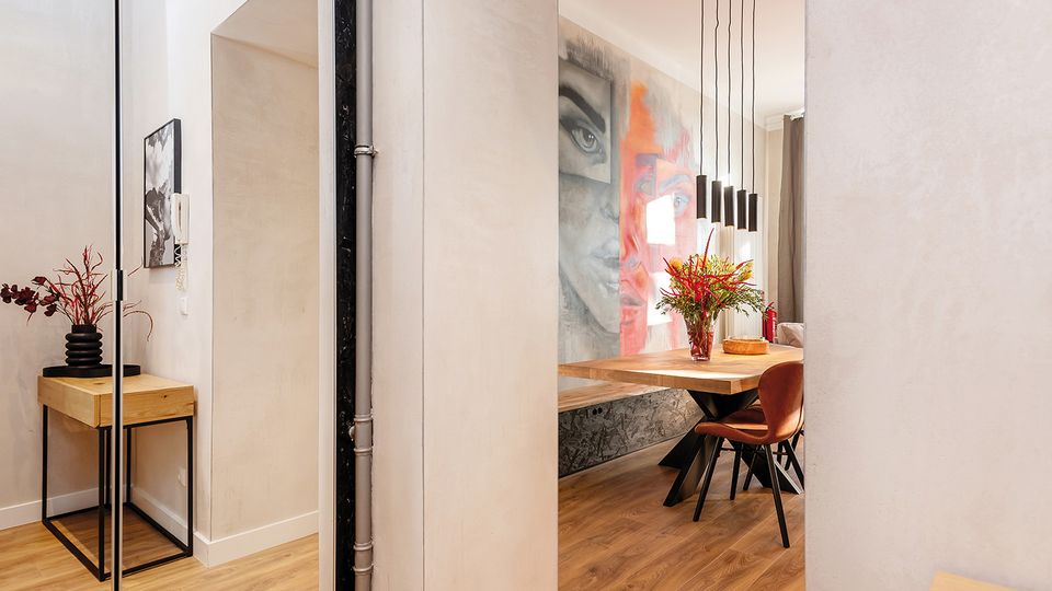 Blick durch einen Flur in einen modernen Essbereich mit Holzboden, großem Holztisch und braunen Stühlen. Über dem Tisch hängen mehrere schwarze Pendelleuchten, an der Wand ist ein großformatiges Wandbild, und seitlich steht eine schmale Konsole mit Vase.