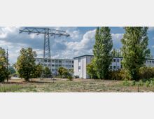 Weitläufige Außenaufnahme eines Industriegeländes mit einem großen Strommast im Vordergrund, Bürogebäuden im Hintergrund und umgebender Vegetation aus Bäumen und Sträuchern. Der Boden ist überwiegend trocken und bewachsen mit spärlichem Gras.