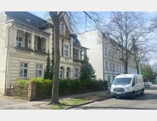Straßenszene mit Altbauhäusern in heller Fassade im Vordergrund, umgeben von kleinen Bäumen und einem Metallzaun. Auf der Straße parkt ein weißer Lieferwagen, und im Hintergrund sind weitere Gebäude und vereinzelte Bäume sichtbar.