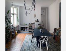 Esszimmer in einer Altbauwohnung mit Holzdielenboden und hoher Decke. Zu sehen sind ein großer dunkler Esstisch mit verschiedenen Stühlen, eine weiße Küche im Hintergrund und eine Fensterfront mit Pflanzen auf der linken Seite.