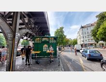 Eine grüne historische Kioskstruktur steht unter einer Eisenbahnbrücke, umgeben von Menschen, die an Holztischen stehen. Im Hintergrund sind eine belebte Straße mit Autos, Baustellenabsperrungen sowie mehrstöckige Wohngebäude und Bäume zu sehen.