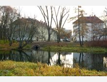 Wohngebäude am Ufer eines kleinen Teichs, umgeben von kahlen Bäumen und Herbstvegetation. Im Vordergrund ist der Teich mit einer Grasuferböschung zu sehen, während im Hintergrund Wohnhäuser in heller Farbe stehen.