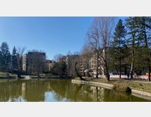 Ein kleiner Park mit einem Teich im Vordergrund, umgeben von Bäumen und mehrstöckigen Wohngebäuden im Hintergrund. Auf der rechten Seite befinden sich Bauabsperrungen und ein Gehweg, der entlang des Teiches verläuft.
