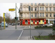 Straßenszene mit einer historischen Eckhausfassade, die im Erdgeschoss ein Lokal mit der Aufschrift „Schultheiss Groß-Destillation Quelle“ zeigt. Im Vordergrund befinden sich eine Straßenkreuzung mit Verkehrsampeln, ein gelbes Schild mit Wegweiser nach „Charlottenburg/Schöneberg“ und ein Eingang zur U-Bahn mit einem blauen „U“-Schild.