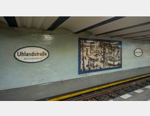 U-Bahnhof Uhlandstraße in Berlin mit hellblau gefliesten Wänden, ovalen Stationsschildern und einem historischen Schwarz-Weiß-Foto, das eine belebte Straßenansicht zeigt. Im Vordergrund sind Gleise mit gelbem Sicherheitsstreifen sichtbar.