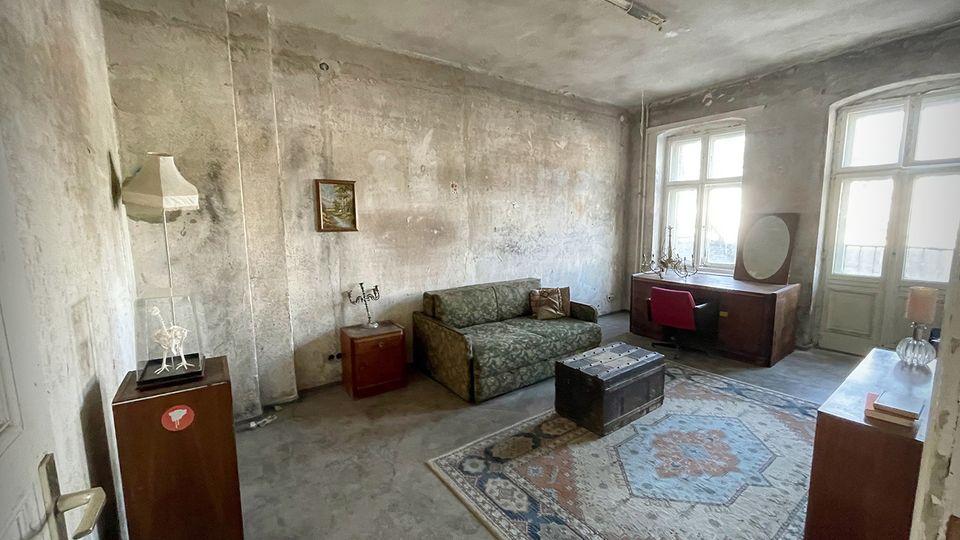 Abgenutztes Wohnzimmer in einem älteren Gebäude mit rohen, fleckigen Wänden und einer einfachen Deckenleuchte. Vor zwei großen Fenstern stehen ein Schreibtisch und eine Kommode; im Raum befinden sich zudem ein gemustertes Sofa, ein Teppich und eine niedrige Truhe als Couchtisch.