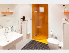 Modernes Badezimmer mit weißen Wandfliesen, schwarzem Fliesenboden und einer ebenerdigen Dusche mit orangenen Fliesen und Glastür. Im Raum befinden sich ein Waschbecken mit Spiegel, ein Haarfön, ein Handtuchhalter sowie eine Toilette und ein Regal mit Handtüchern und Dekoration.