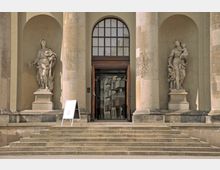 Eingangsbereich eines historischen Gebäudes mit großen Säulen, zwei Nischen mit steinernen Statuen und einer gläsernen Tür in einem Holzeingangsrahmen; eine Treppe führt hinauf zum Eingang. Ein Aufsteller steht links neben der Treppe.