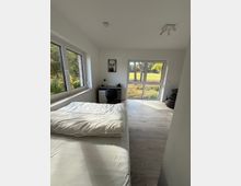 Helles Schlafzimmer mit großem Fenster zur rechten Seite, das den Blick in eine grüne Landschaft ermöglicht. Im Raum befinden sich ein Bett mit weißer Bettwäsche, ein Schreibtisch mit Stuhl sowie ein Bild an der Wand; der Boden ist mit hellem Holzlaminat ausgelegt.