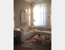 Sunlit living room with a cream sectional sofa, textured throw pillows and blankets, and a low round wooden coffee table on a shag rug. A large window with sheer curtains, a curved floor lamp, and a woven wall hanging decorate the neutral-toned space with wood flooring.
