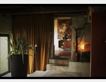 Raum mit rustikalem Interieur, bestehend aus einem großen Topfpflanzengefäß, goldfarbenem Vorhang und einer Treppe, die in einen Nebenraum mit Holzboden führt, wo ein Sideboard und ein großformatiges Blumenbild sichtbar sind. Ein leuchtender Lampenschirm sowie dekorative Elemente wie ein Kürbis und ein dürrer Ast in einer Vase ergänzen die Szenerie.