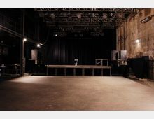 Industriehalle mit einer kleinen Bühne im Vordergrund, beleuchtet durch zahlreiche Scheinwerfer und umgeben von schwarzen Lautsprechern. Die Wände sind roh und wirken unverputzt, während der offene Raum durch dunkle Vorhänge und Metallstrukturen geprägt ist.