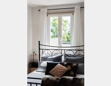 Ein helles Schlafzimmer mit einem Metallbettgestell und mehreren dekorativen Kissen in Grau und Schwarz. Im Hintergrund befindet sich ein großes Fenster mit weißen Vorhängen, durch das Bäume und ein blauer Himmel zu sehen sind.