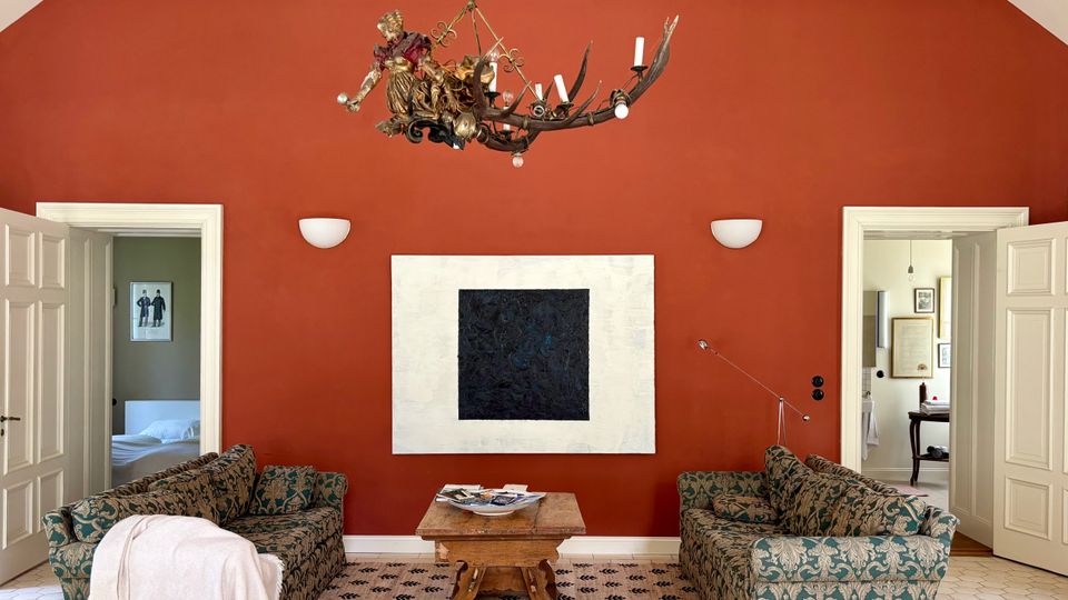Wohnzimmer mit hoher, spitz zulaufender roter Akzentwand, großer abstrakter Wandkunst und dekorativem Kronleuchter. Zwei gemusterte Sofas stehen sich auf einem Teppich gegenüber, dazwischen ein Holztisch; seitlich führen offene weiße Doppeltüren in angrenzende Räume.