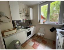 Kleine, helle Küche mit weißen Einbauschränken, Arbeitsplatte mit Herd und Backofen sowie offenem Regal und Edelstahl-Rückwand. Großes Fenster mit Blick ins Grüne, gefliester Boden in Beige- und Terrakottatönen, dazu ein Stuhl neben der Fensterbank und eine Spüle auf der rechten Seite.