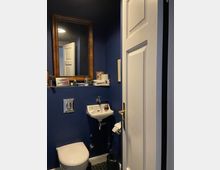 Kleines WC mit dunkelblauen Wänden, wandhängendem Waschbecken und Toilette sowie sechseckigen Bodenfliesen. Über dem Waschbecken hängt ein großer Spiegel mit Holzrahmen und einer schmalen Ablage mit Handtüchern und Pflegeartikeln; eine weiße Tür steht halb offen.