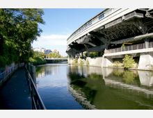 Fußweg entlang eines Kanals mit Geländer auf der linken Seite; rechts eine Hochbahnbrücke aus Stahl, die über das Wasser führt. Im Hintergrund sind Wohngebäude, eine kleine Straßenbrücke und grüne Vegetation sichtbar.