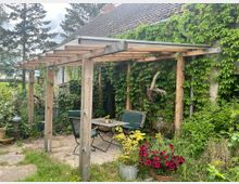 Holzpergola mit transparentem Dach an einem mit Efeu bewachsenen Haus. Darunter befinden sich ein Tisch mit Stühlen und umgebende Pflanzkübel mit bunten Blumen; im Hintergrund sind Bäume und dichtes Grün zu sehen.
