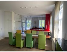 Moderne Wohnküche mit großem Esstisch aus Holz, umgeben von Stühlen mit bunten Stoffbezügen in Grün und Gelb. Im Hintergrund eine Küchenzeile mit roten Hochglanzfronten, großen Fenstern und viel Tageslicht.