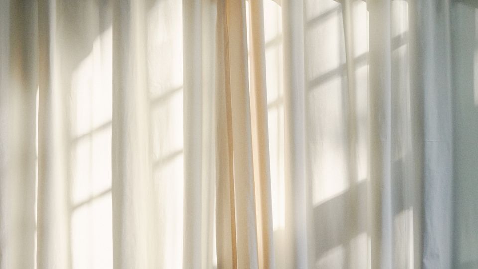Halbtransparente, weiße Vorhänge vor einem Fenster, durch die warmes Sonnenlicht fällt. Die Fenstersprossen zeichnen sich als Schattenmuster auf dem Stoff ab.