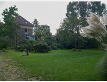 Weitläufiger Garten mit einer großen Rasenfläche, umgeben von Bäumen, Sträuchern und dekorativem Ziergras im Vordergrund; im Hintergrund steht ein zweistöckiges Wohnhaus mit rotem Dach und Wintergarten.