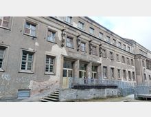 Fassade eines alten Gebäudes mit der Aufschrift „Stadtbad Lichtenberg“. Das Gebäude hat eine graue, teilweise beschädigte Fassade mit mehreren Fenstern, Statuen zwischen den oberen Fenstern und zugänglicher Rampe vor dem Eingang.