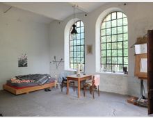 Ein einfacher Raum mit hohen Decken, großen, bogenförmigen Fenstern mit schwarzem Gitterrahmen, die Tageslicht hereinlassen. Der Raum enthält ein Holzbett mit Decken, einen Holztisch mit Stühlen und eine Staffelei; die Wände sind schlicht mit minimaler Dekoration, und der Betonboden wirkt rau.