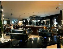 Innenraum eines modernen Restaurants mit gedämpfter Beleuchtung. Im Vordergrund stehen mehrere Warmhaltebehälter auf einem Tisch, während im Hintergrund Gäste an Tischen sitzen und eine Bar mit hängenden Lampen sichtbar ist.