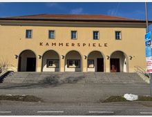 Außenansicht eines Gebäudes mit gelber Fassade und rotem Schriftzug „KAMMERSPIELE“ über fünf Rundbögen. Vor dem Eingang führt eine breite Treppe hinauf, daneben befindet sich eine gepflasterte Fläche.