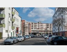 Straßenszene mit Wohngebäuden auf beiden Seiten und einem mehrstöckigen Gebäude im Hintergrund mit Balkonen und Glasfassade im Erdgeschoss. Autos sind auf beiden Seiten der Straße geparkt, Bäume und Laternen säumen den Weg, und einige Passanten sind sichtbar.